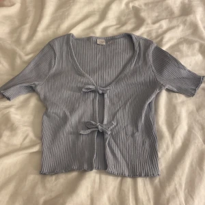 Ljuslblå ribbad topp från Lindex - Superfin ljusblå ribbad topp från Lindex med korta ärmar och två söta knyt framtill. Perfekt att styla med höga jeans eller kjol. Mjuk och stretchig i materialet för en bekväm känsla.