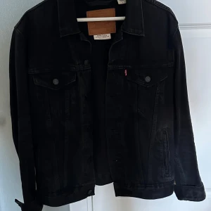 Svart jeansjacka från Levi's  - Säljer en svart jeansjacka från Levi's Premium i klassisk modell. Jackan har knappar framtill, två bröstfickor med lock och knapp samt sidofickor. Snygg, tidlös och passar perfekt till lager-på-lager. Materialet är robust denim och jackan har lång ärm.