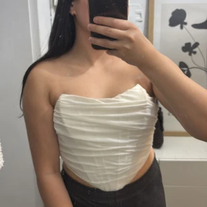 Vit korsett med dragkedja - Säljer en snygg vit korsett med draperad effekt och markerad midja. Toppen är axelbandslös och har en silkeslen känsla med dragkedja bak för enkel påklädning. Perfekt för dig som vill sticka ut med en elegant och trendig look.