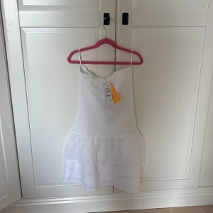 Vit smockklänning från Gina Tricot - Helt oanvänd supersöt vit smockklänning från Gina Tricot med volang i nederdelen. Klänningen har en smockad överdel och är kort i modellen, perfekt för sommarens alla tillfällen. Materialet är mjukt och lätt, och volangen ger en extra feminin touch.