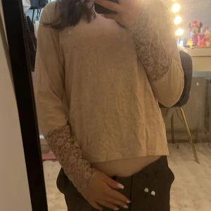 Beige långärmad topp med spetsdetaljer - Säljer en beige långärmad topp med snygga spetsdetaljer på ärmarna. Spetsen har ett blommigt mönster som ger en romantisk och trendig vibe. 