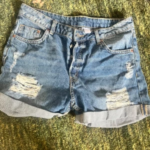 Blå jeansshorts med slitningar - Snygga blå jeansshorts med slitna detaljer och upprullade benslut. Klassisk femficksmodell med dragkedja och knapp framtill. Perfekta för sommaren och ger en avslappnad vibe. Passar dig som gillar en lite loose fit och streetstyle.