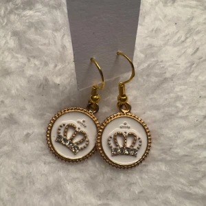 Guldiga örhängen med kronmotiv - Snygga örhängen i guld med rund vit platta och glittrande krona i mitten. Perfekta för dig som vill sticka ut med en kunglig touch. Hängande modell med fina detaljer runt kanten. Krokarna är i sterling silver