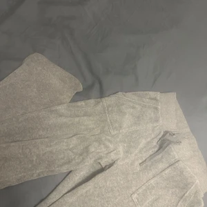 Grå mjukisbyxor med fickor och snörning - Säljer ett par grå mjukisbyxor med bred resår i midjan och snörning framtill. Byxorna har två stora fickor med lock och knapp bak, samt mjukt material som är perfekt för chill eller träning.