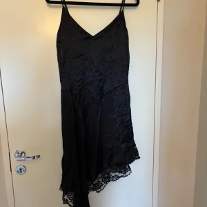 Svart satin klänning med spetsdetaljer - Säljer en svart klänning i satin med v-ringning och smala axelband. Klänningen har asymmetrisk nederkant med fina spetsdetaljer. Perfekt för dig som vill ha en elegant och stilren look.