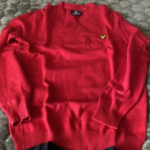 Röd tröja från Lyle & Scott - Säljer en klassisk röd tröja från Lyle & Scott med rund halsringning och den ikoniska gula loggan broderad på bröstet. Tröjan har långa ärmar och ribbade muddar vid ärmslut och nederkant. Perfekt för lager-på-lager-stil. helt ny