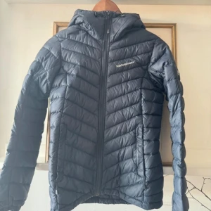 Marinblå  pufferjacka från Peak Performance - Säljer en marinblå pufferjacka från Peak Performance med huva och dragkedja framtill. Jackan har en sportig look och är lätt vadderad, perfekt för kyliga dagar. Snygg och enkel design med logga på bröstet.