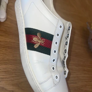 Gucci Ace sneakers med bi-detalj - Säljer ett par vita Gucci Ace sneakers med klassiska gröna och röda ränder samt broderad guldfärgad bi på sidan. Baksidan har metallicröd och metallicgrön detalj med Gucci-logga. Skorna är i skinn och kommer med originalförpackning och dustbag. Och kvitto finns.