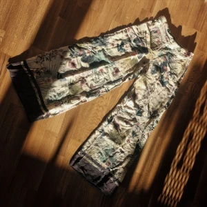 Blommiga vida linnebyxor - Säljer ett par luftiga och vida linnebyxor med blommigt mönster i grönt, lila och beige. Byxorna har mörka kanter nertill och hög midja. Perfekta för varma dagar och ger en avslappnad men stilren look.