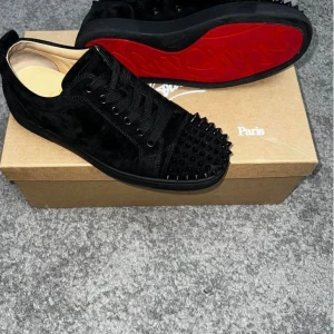 Svarta sneakers med nitar och röd sula - Säljer ett par svarta sneakers med coola nitar på tån och klassisk röd sula. Skorna har snörning och är tillverkade i mocka med platt sula. Perfekta för dig som vill sticka ut med din stil.