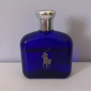 Blå parfymflaska från Ralph Lauren Polo - Säljer en snygg parfymflaska från Ralph Lauren Polo med klassisk blå glasdesign och silverfärgat lock. Flaskan har den ikoniska polospelaren i silver på framsidan. Perfekt för dig som gillar stilrena och sportiga dofter.