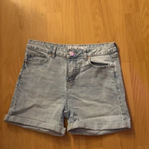 Ljusblå jeansshorts med upprullad kant - Ljusblå jeansshorts med klassisk femficksdesign och upprullad kant. Shortsens passform är normal och de har bälteshällor samt knappstängning framtill. Perfekta för varma dagar.