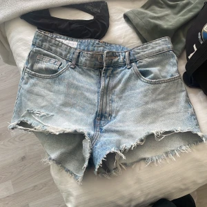 Ljusblå jeansshorts med fransar - Säljer ett par ljusblå jeansshorts med råa franskantade benslut och klassisk femficksdesign. Shortsen har normal passform och är perfekta för varma dagar. Stängs med knapp och dragkedja framtill.