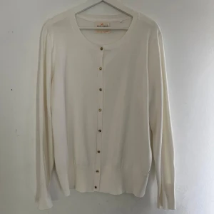 Vit kofta med guldknappar - Säljer en klassisk vit kofta med rund halsringning och guldknappar framtill. Tröjan har långa ärmar och ribbade muddar vid ärmslut och nederkant. Har en fläck på nederdelen på ena armen (se bild 3)
