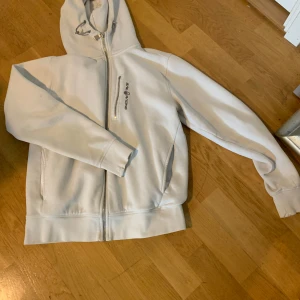 Vit hoodie med dragkedja från Peak Performance - Vit hoodie från Peak Performance med dragkedja framtill och på bröstfickan. Klassisk huva och två sidofickor. Perfekt för en avslappnad stil och enkel att matcha med det mesta.