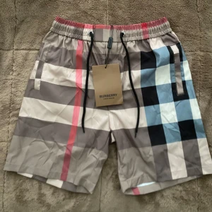 Burberry rutiga shorts med snörning - Snygga shorts från Burberry med klassiskt rutigt mönster i beige, svart, vitt, blått och rött. Shortsen har elastisk midja med svart snörning och sidofickor. Perfekta för sommaren och ger en exklusiv känsla till din outfit. Skriv vid funderingar. Str M