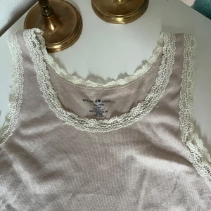 Linne från brandy  - Säljer en söt beige linnetopp från Brandy med fina vita spetsdetaljer längs urringning och ärmhål. Toppen är ärmlös och har en mjuk, bekväm känsla – perfekt för varma dagar eller som lager under en kofta.