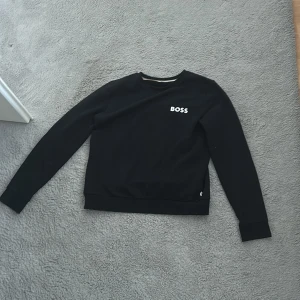 Svart sweatshirt från BOSS - Säljer en svart sweatshirt från BOSS med rund halsringning och diskret logga på bröstet. Tröjan har långa ärmar och ribbade muddar vid ärmslut och nederkant. Perfekt för en stilren och enkel look. Och är bara använd 2 gånger.