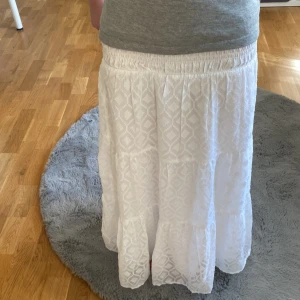 Vit långkjol med mönster - Säljer en vit långkjol från Gina Tricot med diskret geometriskt mönster och bred resår i midjan. Kjolen är luftig och har flera volanger för extra rörelse. Perfekt för varma dagar och enkel att matcha med olika toppar.