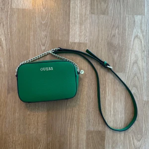 Grön axelväska från Guess - Snygg grön axelväska från Guess med silverfärgad kedja och justerbar axelrem. Väskan har ett stilrent utseende med Guess-logga framtill och dragkedja upptill. Perfekt för att lyfta din outfit. Aldrig använd. 