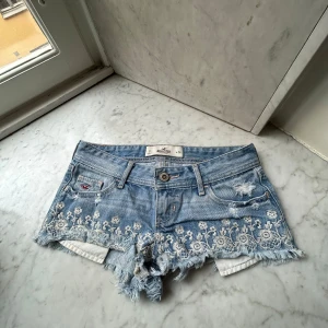 Hollister shorts - Säljer ett par ljusblå jeansshorts från Hollister med slitna kanter och vitt blommigt broderi framtill. Klassisk femficksmodell med knapp och dragkedja. Perfekta för sommaren!
