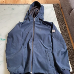 Marinblå jacka med huva från Sail Racing - Säljer en marinblå jacka från Sail Racing med huva och dragkedja. Jackan har diskret logotyp på bröstet och en rund Sail Racing-detalj på ärmen. Insidan är fodrad med mjuk fleece för extra komfort. Perfekt för blåsiga dagar.