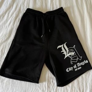 Svarta shorts med LA-tryck - Feta svarta shorts med vit LA-grafik och texten 'City of Angels EST 1850' på ena benet. Resår i midjan med snörning för justerbar passform. Perfekta för en avslappnad streetwear-look.