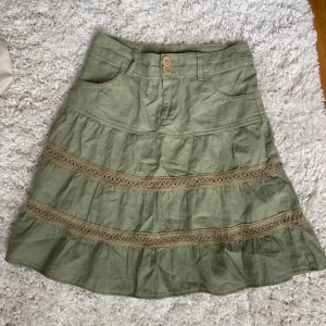 Grön kjol med volang och spetsdetaljer - Säljer en grön kjol med två dekorativa spetsband och volang. Kjolen har fickor fram och stängs med knapp och dragkedja. Perfekt för dig som gillar en bohemisk stil.
