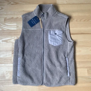 Beige fleeceväst med bröstficka - Mysig beige fleeceväst från Burton med hel dragkedja och kontrasterande bröstficka i ljusgrått tyg. Västen har ståkrage och två sidofickor. Perfekt för lager-på-lager och stilren look.