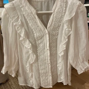 Vit blus med spets och volang från Reserved - Säljer en  vit blus från Reserved med fina spetsdetaljer längs knäppningen och volanger framtill. Blusen har trekvartsärm och knappar framtill. 