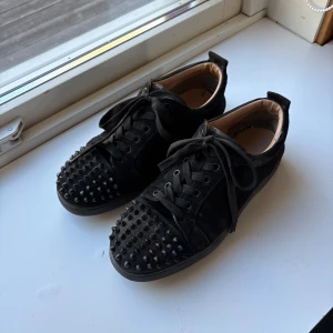 Svarta sneakers med nitar från Christian Louboutin - Säljer ett par svarta sneakers från Christian Louboutin med svarta nitar över hela tån och klassisk röd sula. Skorna har snörning och är tillverkade i mocka med läderdetaljer. Perfekta för dig som vill sticka ut med en edgy stil.