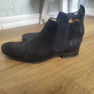 Svarta chelsea boots i mocka från Novita - Stilrena svarta chelsea boots i mocka från Novita. Skorna har elastiska paneler på sidorna och dragflikar bak för enkel påtagning. Nypris är över 2000kr