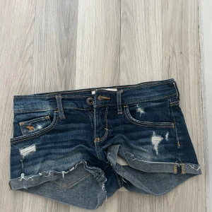 Mörkblå jeansshorts från Abercrombie & Fitch - Snygga mörkblå jeansshorts med slitna detaljer och uppvikta kanter. Klassisk femficksmodell med orangea sömmar och broderad logga på myntfickan. Perfekta för sommaren.