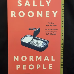 Normal People - Romanen 'Normal People' av Sally Rooney utforskar relationen mellan Connell och Marianne i en småstad på Irland. Boken handlar om vänskap, kärlek och identitet, och följer deras liv från tonåren till vuxen ålder. Omslaget är korallfärgat med illustration.