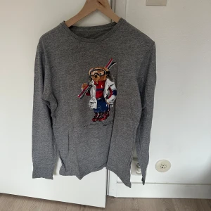 Grå långärmad tröja med björntryck från Polo Ralph Lauren - Säljer en grå långärmad tröja från Polo Ralph Lauren med ett stort tryck av deras ikoniska björn på framsidan. Tröjan har rund halsringning och är i mjuk bomull. Perfekt för en avslappnad och stilren look.
