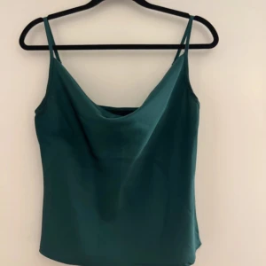 Mörkgrön satin linnetopp - Säljer en elegant mörkgrön linnetopp i satin med smala axelband och snygg draperad urringning. Perfekt att styla till både jeans och kjol för en trendig look.