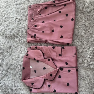 Pyjamas set + tofflor - Ett rosa gulligt pyjamas set med svarta hjärtan. Skjorta med korta ärmar och långa byxor. Samt ett par vita fluffiga tofflor med gulldiga hjärtan. Storlek S. aldrig använda 