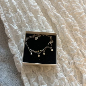 Silverfärgat armband med hängande stenar - Säljer ett snyggt silverfärgat armband med flera små hängande stenar som ger ett glittrigt intryck. Armbandet har justerbar kedja och passar perfekt som accessoar till olika tillfällen.
