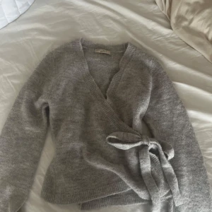 Grå omlottkofta med knytband - Supermjuk och fluffig grå omlottkofta . Koftan har långa ärmar, v-ringning och knytband i midjan för en snygg passform. Perfekt att slänga på sig när det är kyligt ute.