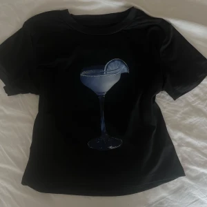 Svart t-shirt med drinkmotiv - Svart t-shirt med ett tryck av ett cocktailglas och lime på bröstet. T-shirten har rund hals och korta ärmar, perfekt för en chill och stilren look. Skönt luftigt material, sitter som storlek s/xs