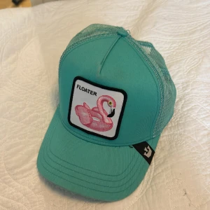 Turkos keps med flamingo från The Farm Goorin Bros - Säljer en turkos keps med mesh-bakstycke och patch framtill med en rosa flamingo och texten 'FLOATER'. Justerbar baktill och märkt med The Farm på insidan.