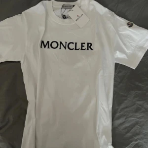 Vit t-shirt från Moncler  - Säljer en vit t-shirt från Moncler med svart Moncler-tryck på bröstet och klassisk logotyp-patch på ärmen. Enkel och stilren design, perfekt för dig som gillar exklusiva märken. Skick 9/10 Storlek M men passar 168-179