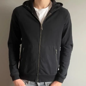 Svart huvtröja från Prada - Snygg svart huvtröja från Prada med dragkedja framtill och två fickor med dragkedjor. Tröjan har en klassisk design med huva och ribbade muddar vid ärmslut och nederkant. Perfekt för en stilren look.