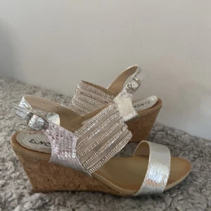 Klackar  - Säljer ett par snygga klackar från Encor med silverfärgade remmar och kilklack i kork. Skorna har öppen tå och justerbart spänne vid ankeln. Perfekta för sommaren och ger en glittrig touch till din outfit.