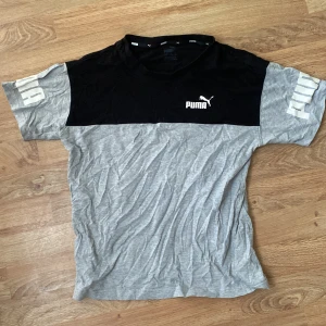 Grå och svart t-shirt från Puma - Säljer en snygg t-shirt från Puma i grått och svart med logga på bröstet och tryck på ärmarna. T-shirten har normal passform och är kortärmad, perfekt för en sportig look.