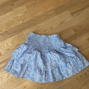 Blå blommig volangkjol från Gina Tricot - Supersöt blå kjol från Gina Tricot med vitt blommönster och bred smock i midjan. Kjolen har volanger och är perfekt för en somrig look. Lätt och luftig modell som är enkel att matcha med olika toppar.