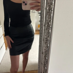 Svart kort kjol i skinnimitation från New Look - Säljer en svart kort kjol i skinnimitation från New Look. Kjolen har en tight passform och är perfekt för dig som gillar en stilren och edgy look. Enkel design utan synliga detaljer, passar till många olika outfits.