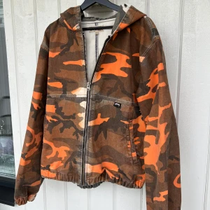 Stüssy camojacka med huva - Säljer en camouflagemönstrad jacka från Stüssy som är ganska rare. Jackan är storlek L men passar passar perfekt för mig som är 175cm lång. Den är gansak tung, dvs bra kvaliten. Enda deffekten är att den har rivits längst lapppen som man ser på bild 4.