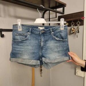 Blå jeansshorts från H&M - Klassiska blå jeansshorts i storlek 40 från H&M med upprullade benslut och femficksmodell. Shortsen har normal passform och är tillverkade i denim med en snygg tvättad look. Perfekta för varma dagar och enkel att matcha med olika toppar.