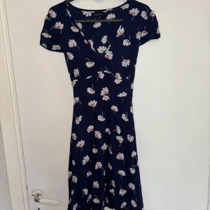 Mörkblå blommig klänning - En söt mörkblå klänning från New Look med vita och orange blommor, v-ringning och korta ärmar. Klänningen har ett knytband i midjan på baksidan. Passar storleken (36/38). Perfekt för sommaren! Fråga gärna för fler bilder! 💙🌸
